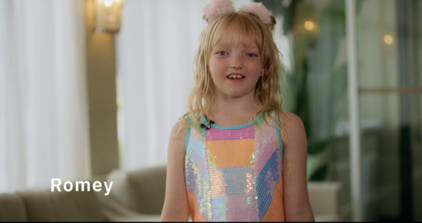 Young girl with blonde hair wearing a colorful sequin dress and a fuzzy headband, smiling indoors with blurred background. "Romey" written on the left.
